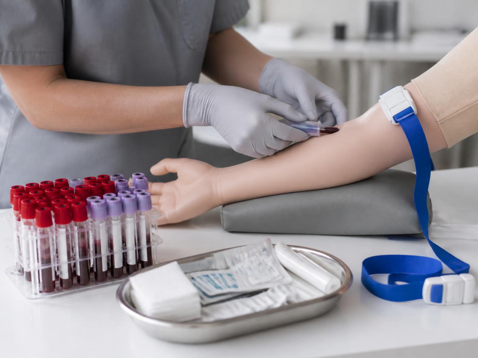 Phlebotomy training: gloved clinician performing venepuncture beside racked blood collection tubes